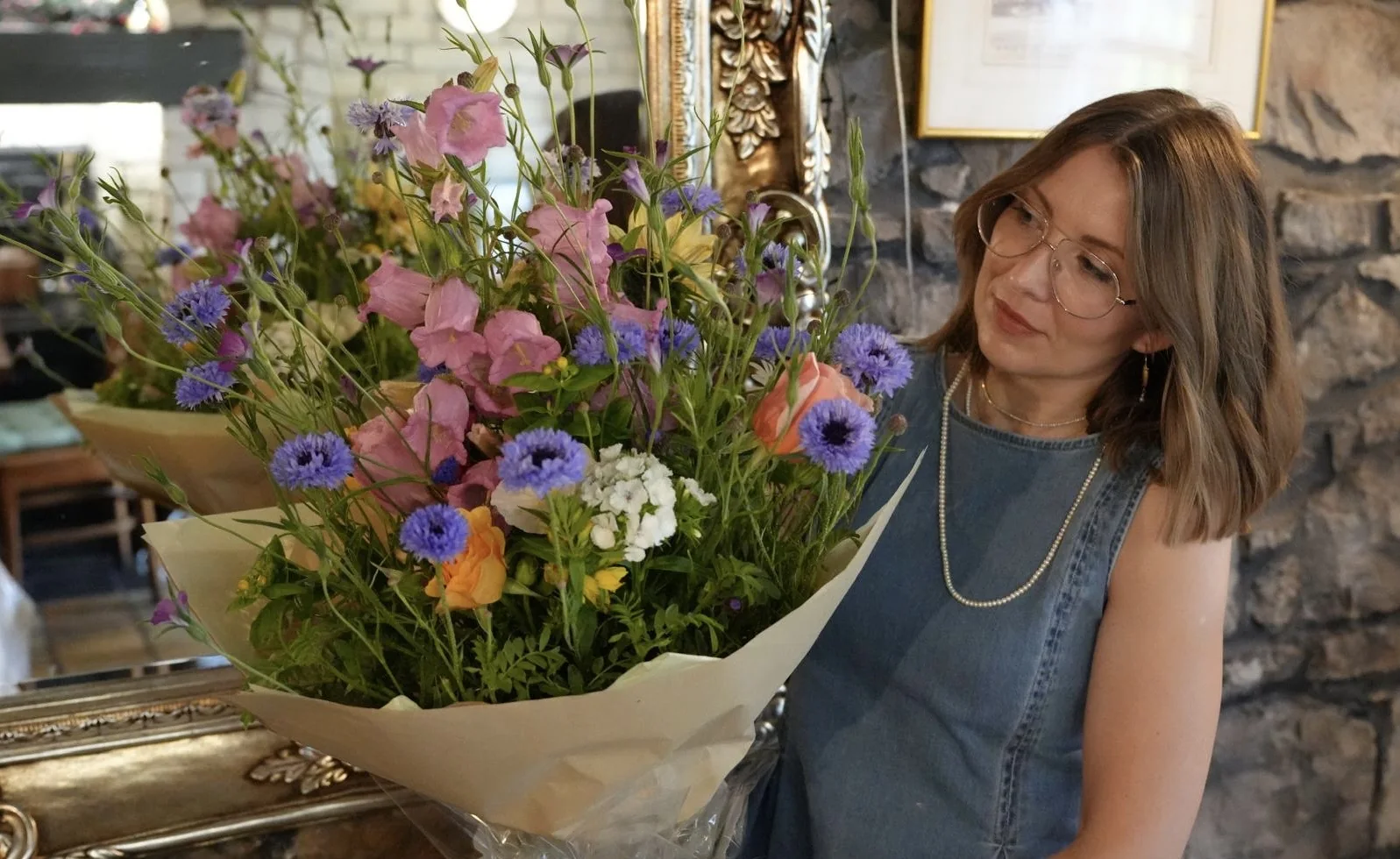 The Cow Shed florist admiring a wrapped bouquet of cornflowers, campanula, roses and sweet williams beside an old stone wall