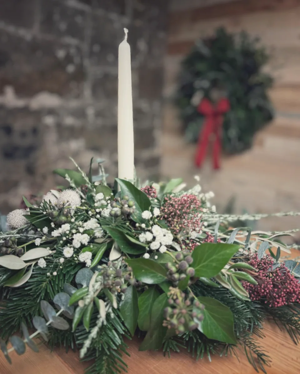 Festive table centrepiece of fir, ivy berries, gypsophila, skimmia and eucalyptus around an ivory taper candle, with a wreath and red bow behind