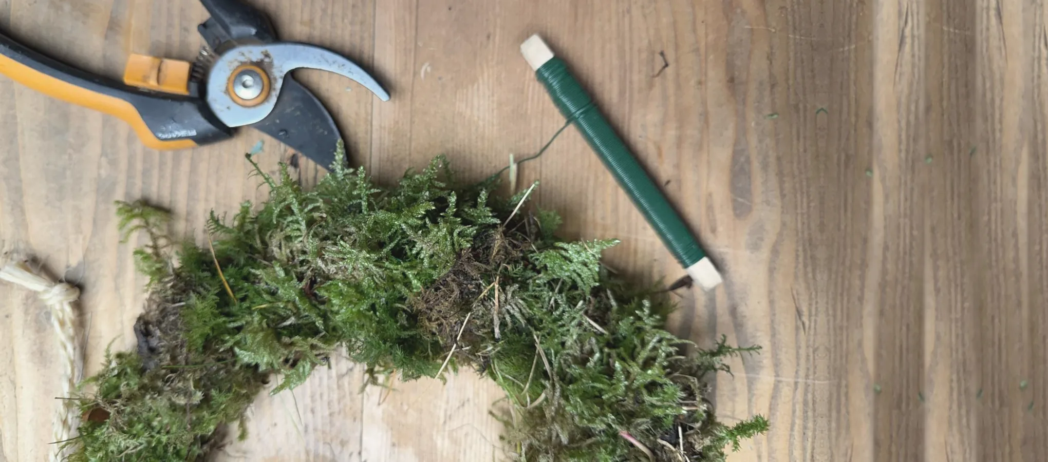Wreath-making workshop table: a moss wreath base, secateurs and a reel of green florist wire on a wooden bench